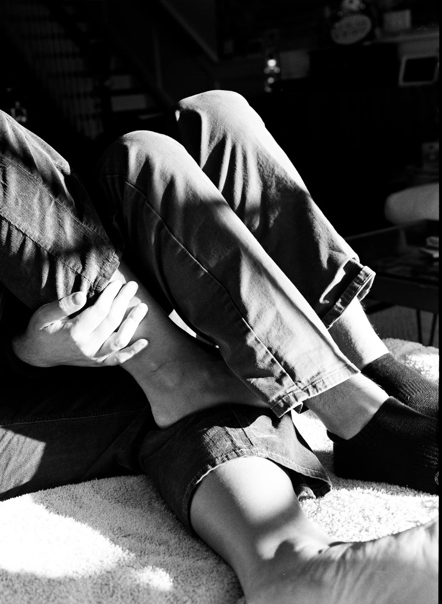 A person is sitting on a carpeted floor, holding their foot and leg while their other leg is bent with their foot flat on the floor. The lighting creates shadows, emphasizing the contours of their legs and hand.