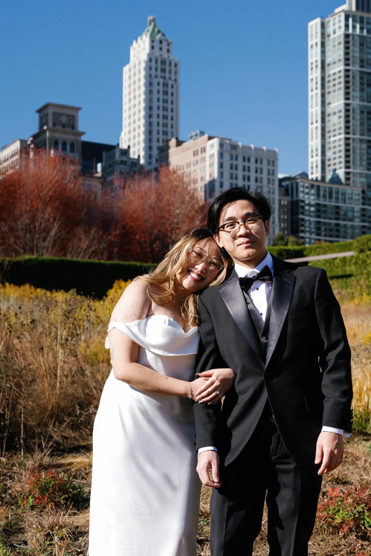 A happy couple dressed in wedding attire, standing outdoors in a park with city skyscrapers in the background, smiling and hugging.