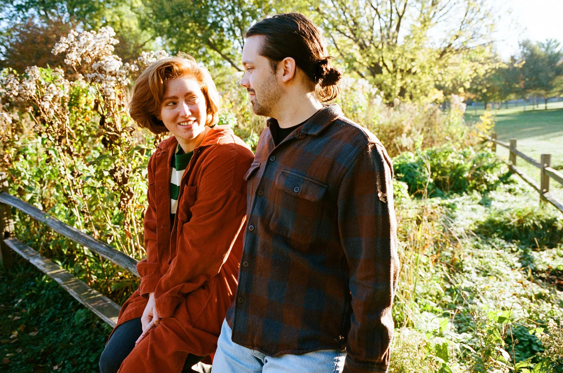 A young man and woman sitting on a park bench outdoors, smiling and looking at each other during sunny autumn day with trees and greenery in the background.