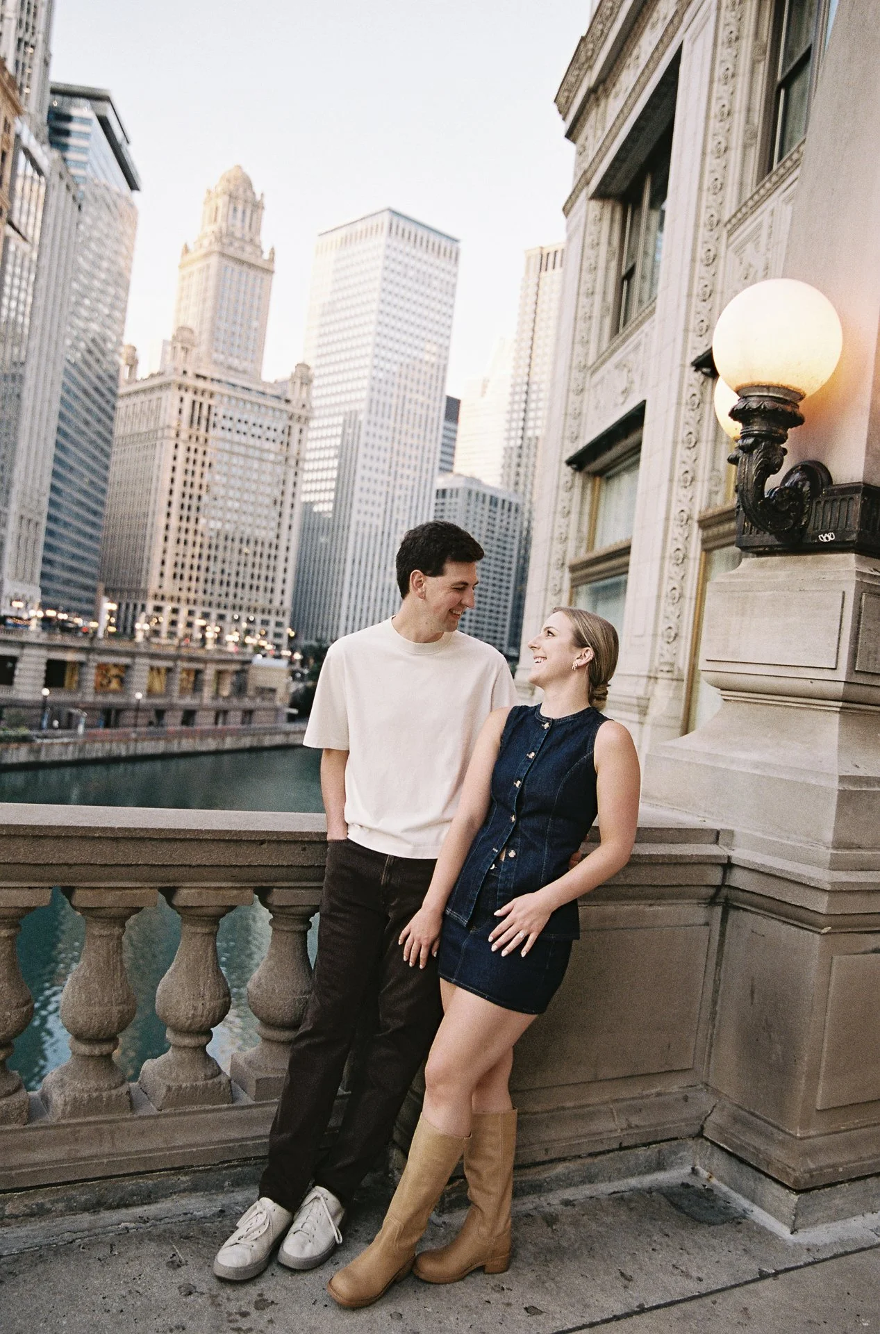 A young man and woman leaning on a stone railing by a river in a city with tall skyscrapers in the background, smiling and looking at each other.