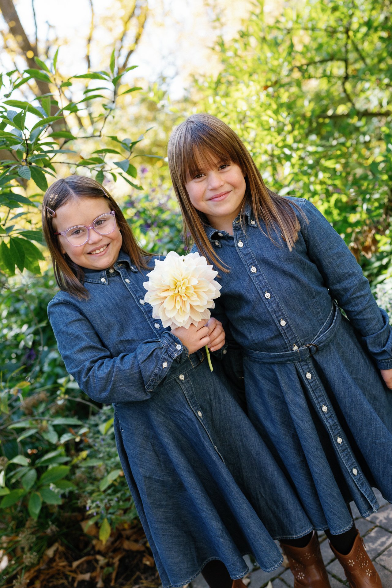 Two young girls in denim dresses smiling outdoors, one girl holding a large light-colored flower, surrounded by green foliage.