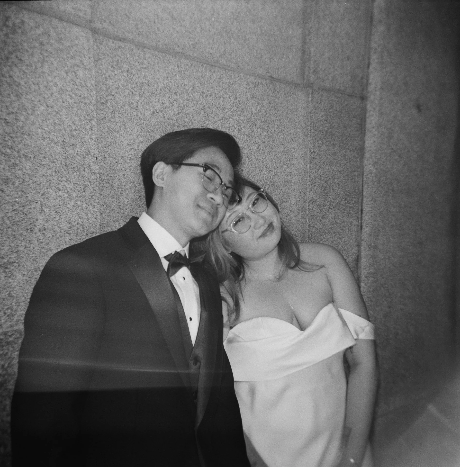 A black and white photo of a young man in a tuxedo and a young woman in an off-shoulder dress leaning against a textured wall.
