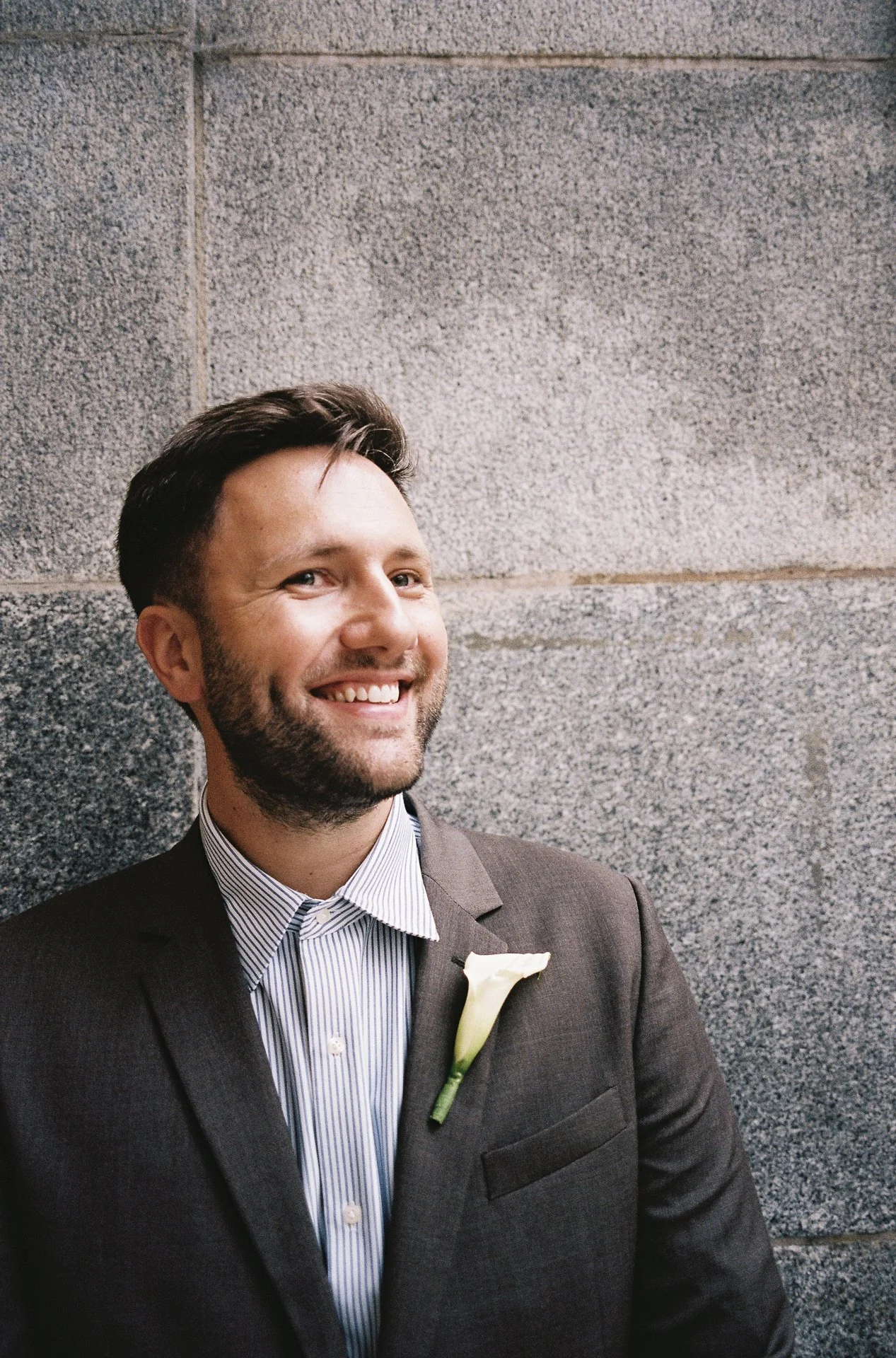 A smiling man in a gray suit with a white flower boutonniere standing against a gray stone wall.