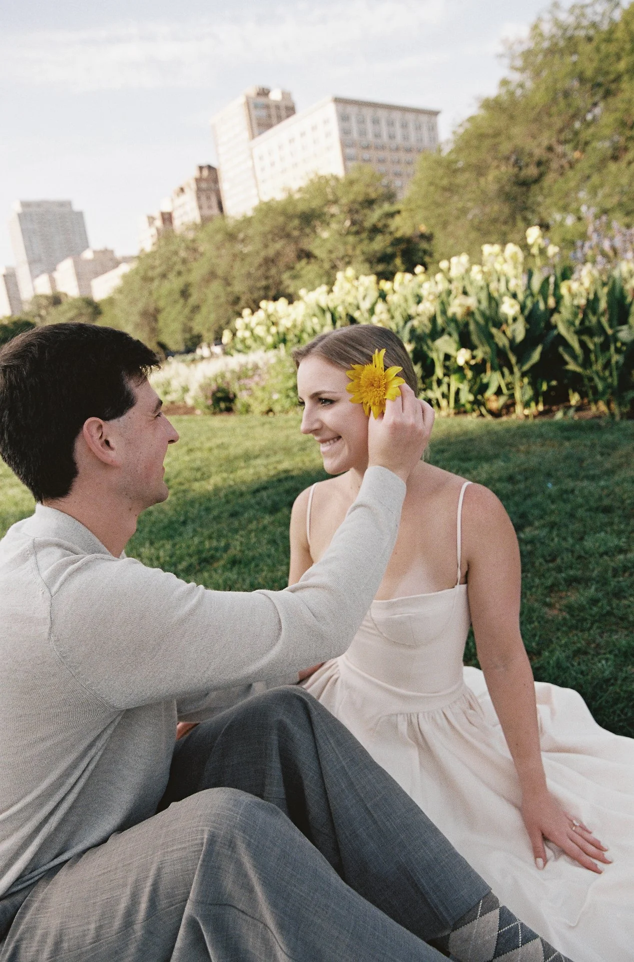 A man holding a yellow flower to a woman's face as they sit on grass in a park with flowers and city buildings in the background.