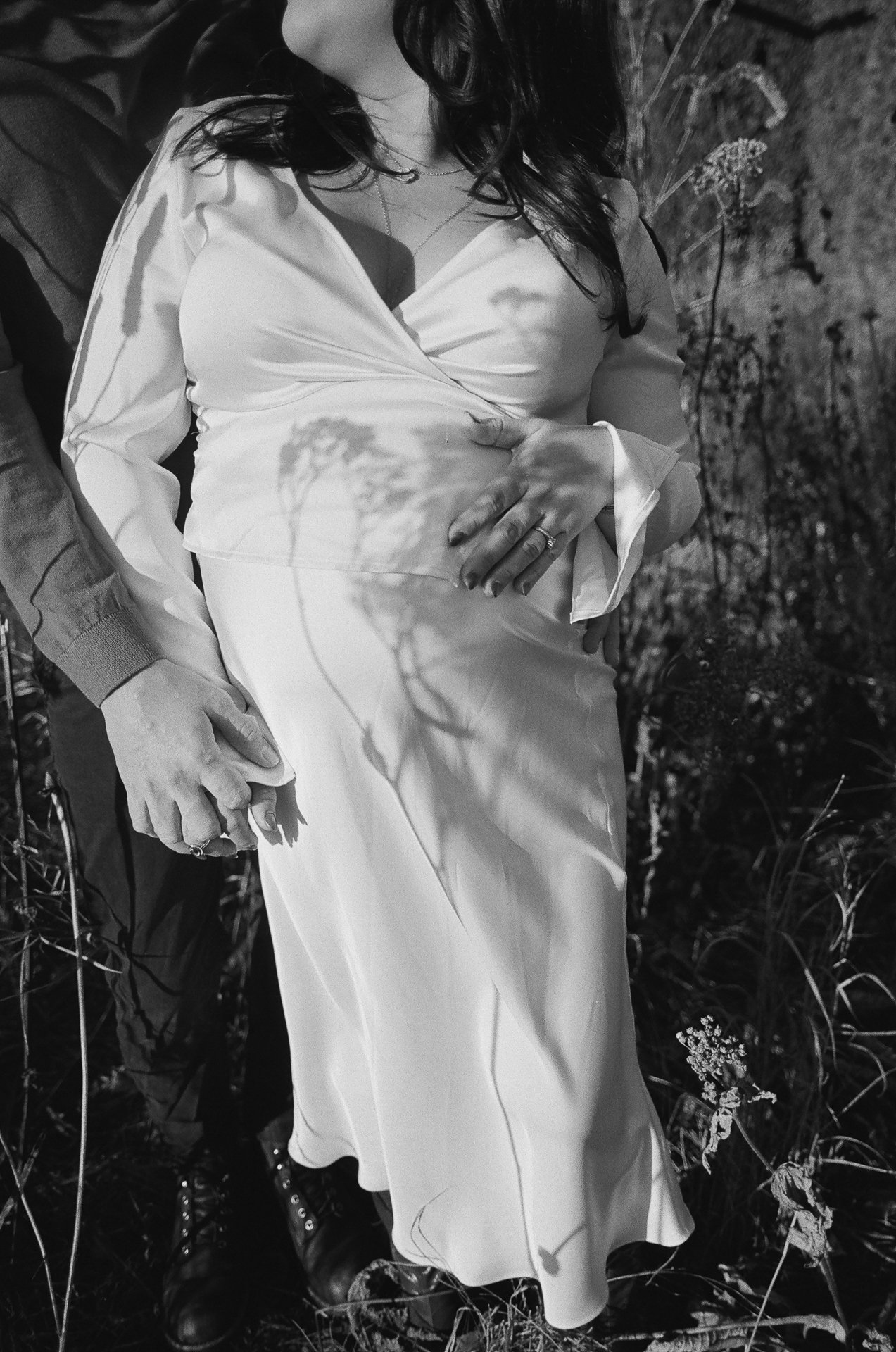 A pregnant woman in a white dress is surrounded by tall grass and plants, with a shadow of a plant cast on her belly. Someone is holding her hand and a shadow of a plant is also on her hand.