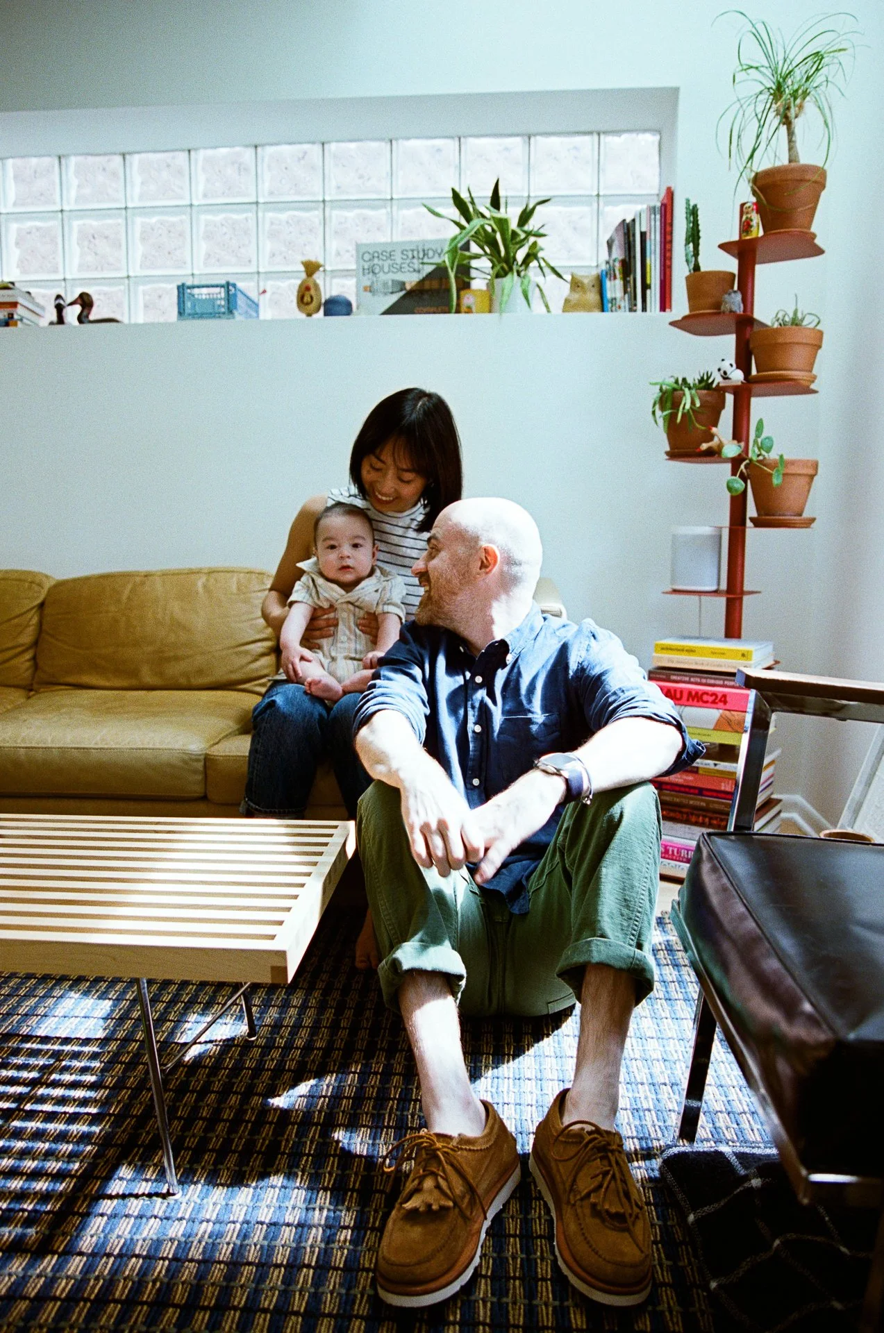 A family sitting in a living room, smiling and interacting. A woman holding a baby on a sofa, a man sitting on the floor, and a bookshelf with potted plants in the background.