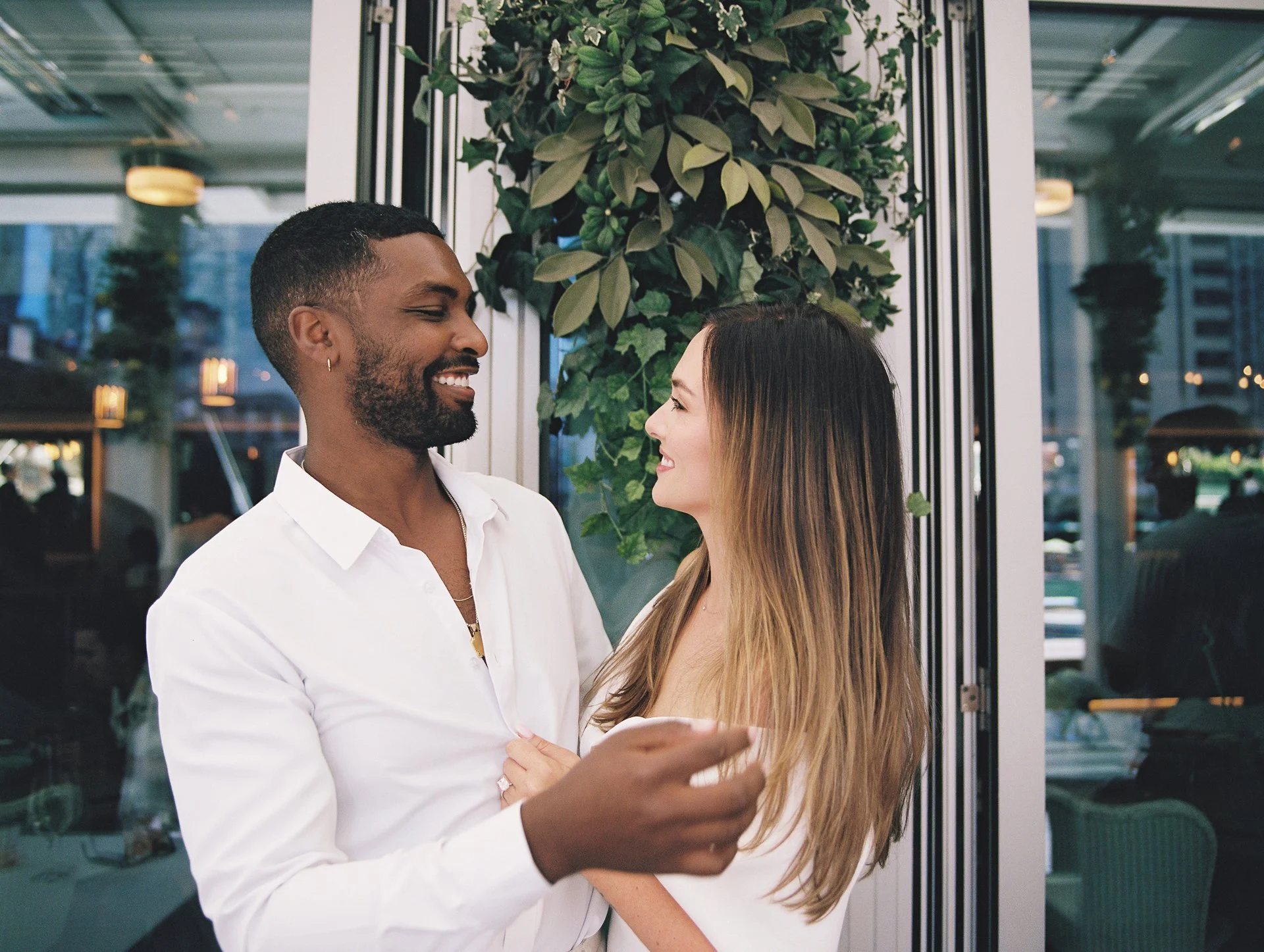 A happy interracial couple dancing and smiling together inside a building with a glass wall and greenery.
