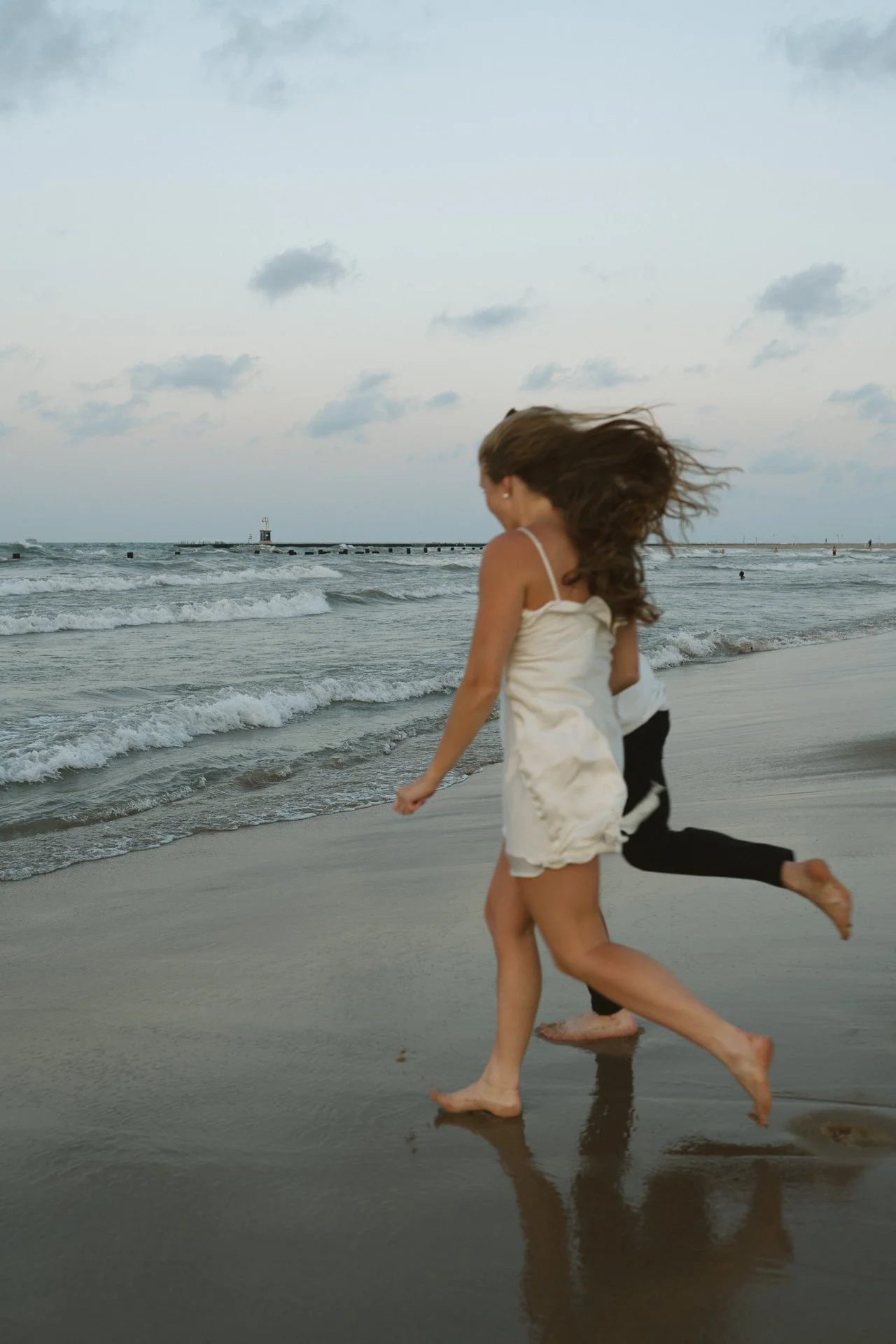 Two women walking along the beach shoreline with the ocean waves rolling in, one wearing a white dress and the other in black pants, with a cloudy sky overhead.