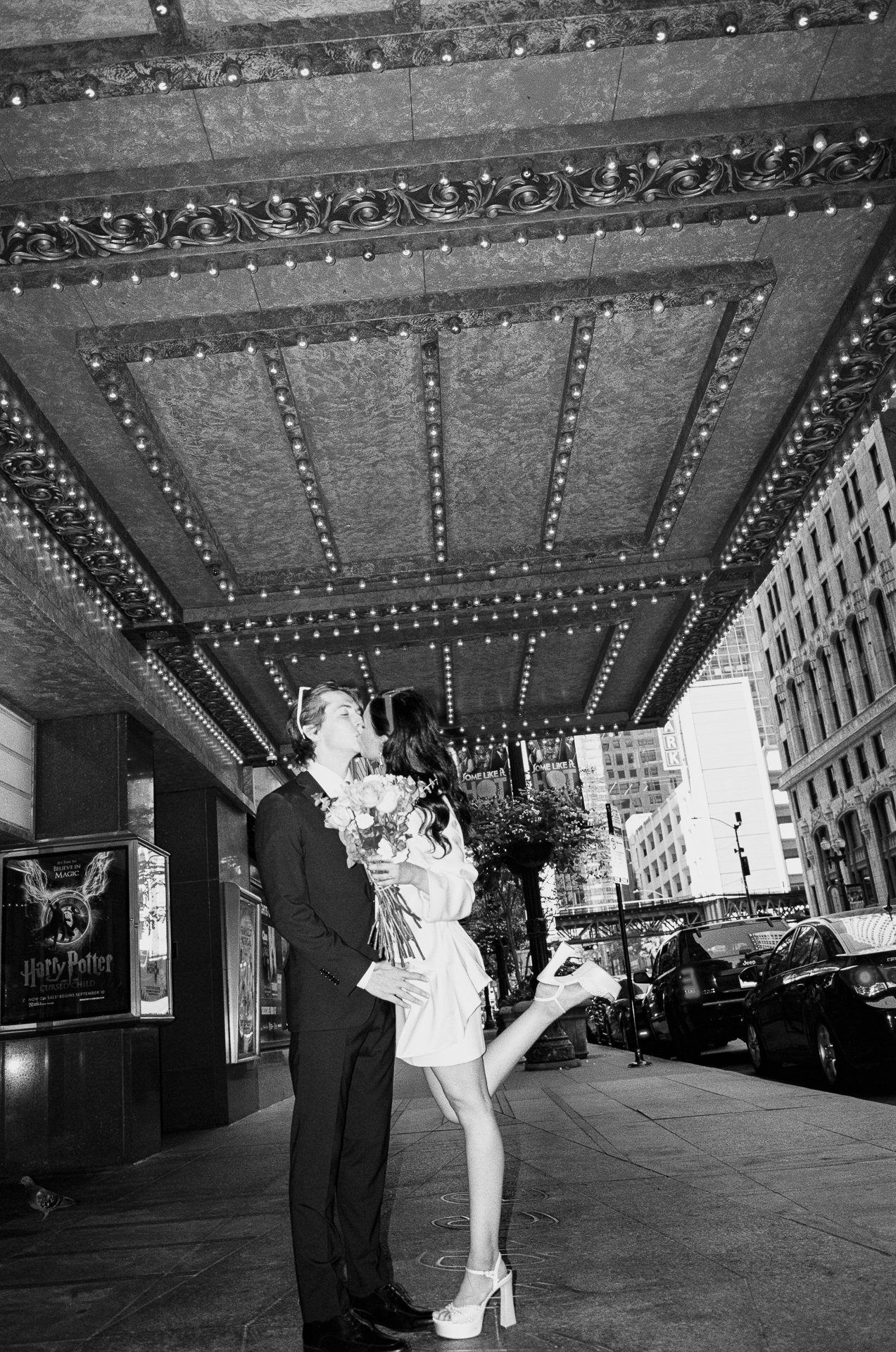 A couple sharing a kiss under a marquee, with the woman holding a bouquet of flowers, on a city sidewalk with tall buildings and parked cars.