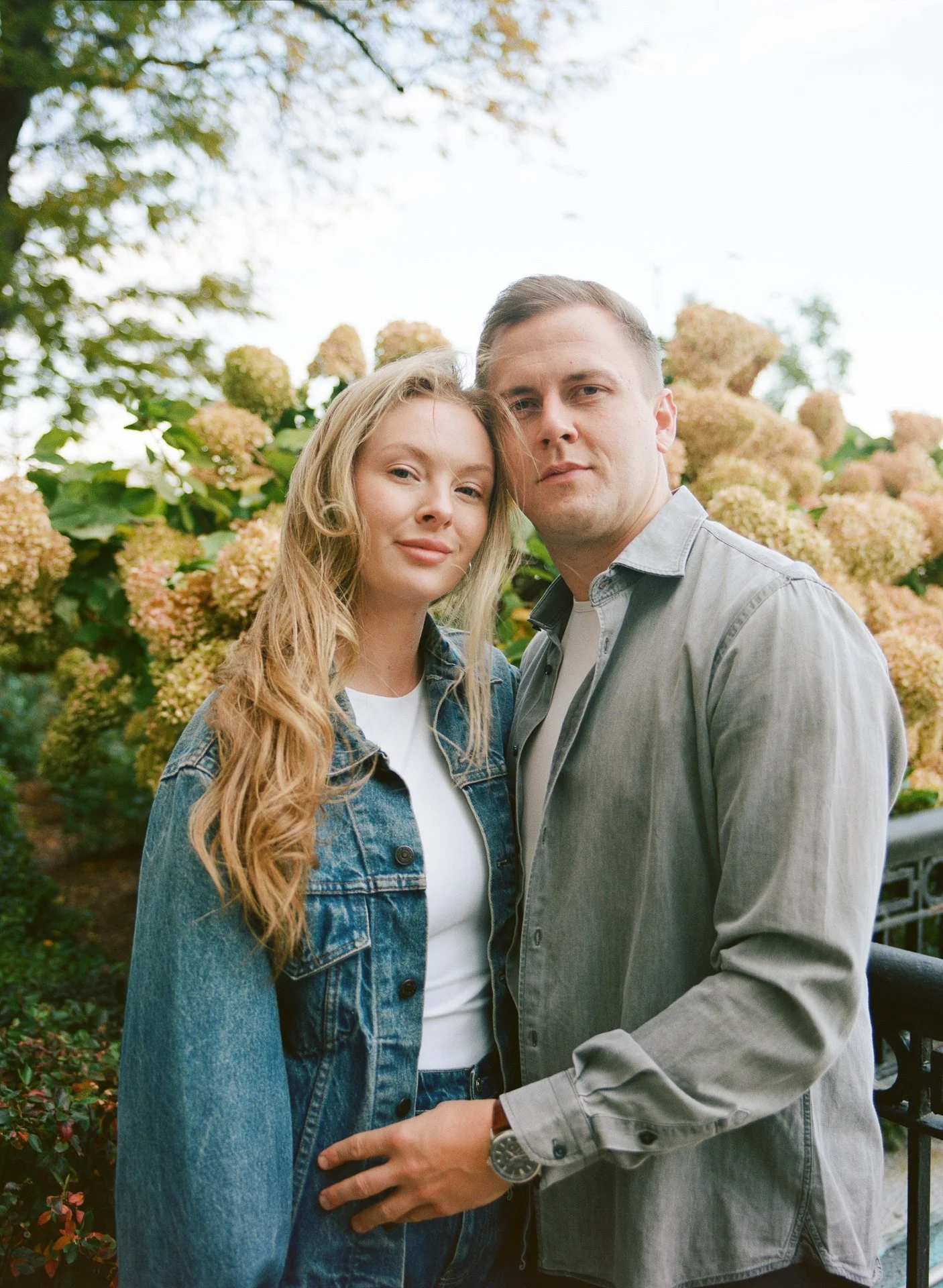 A young woman with long blonde hair in a denim jacket and a young man with short hair in a gray shirt stand close together outdoors, surrounded by lush green foliage and hydrangea flowers.
