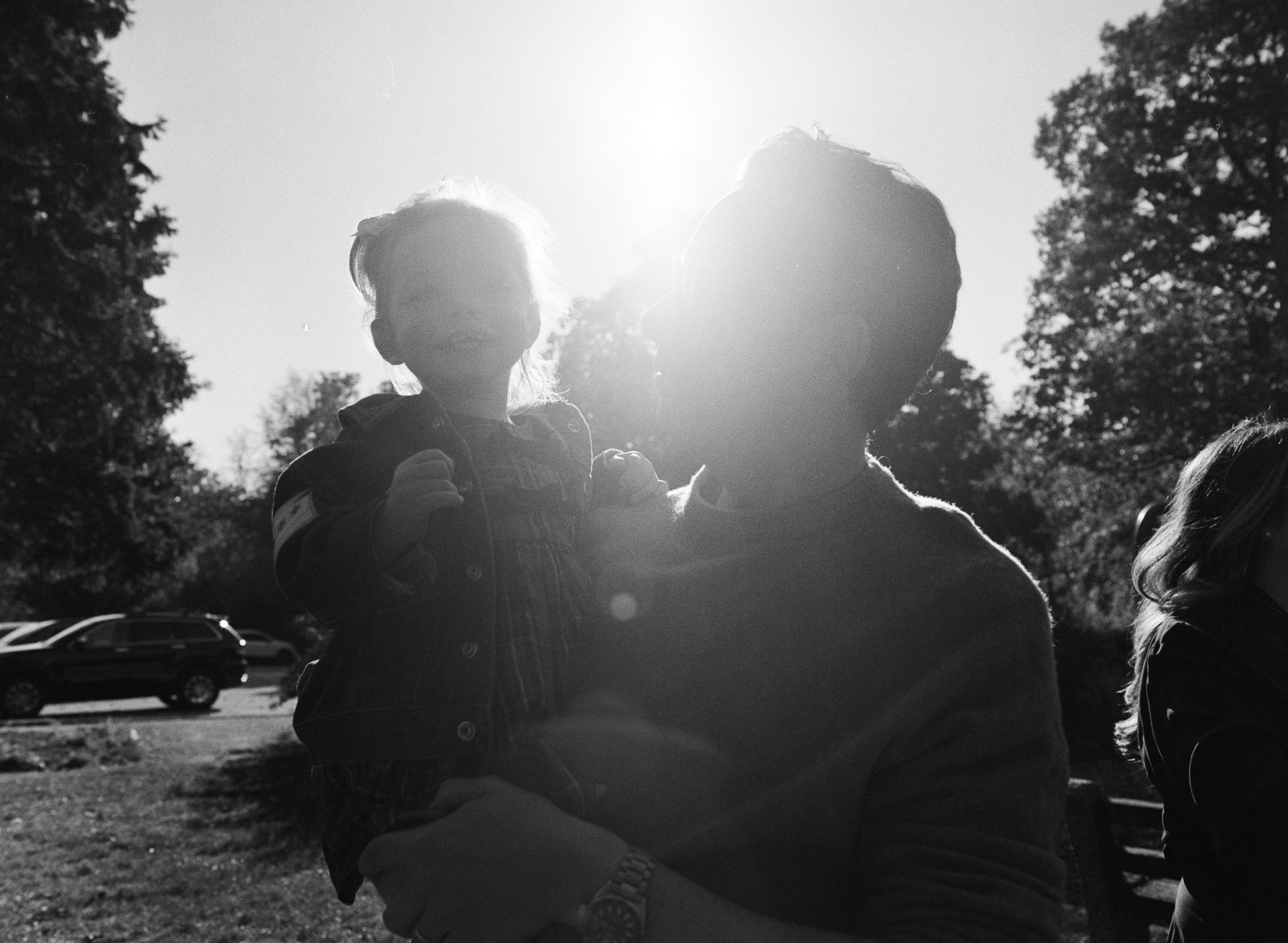 A man holding a young girl outdoors in sunlight, with cars and trees in the background.