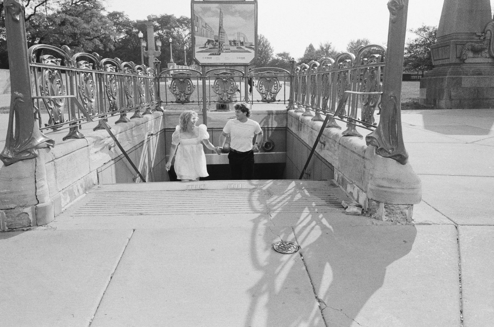 A man and woman holding hands walking up the stairs of a subway entrance, with ornate metal railings and a sign overhead, in a city park setting.