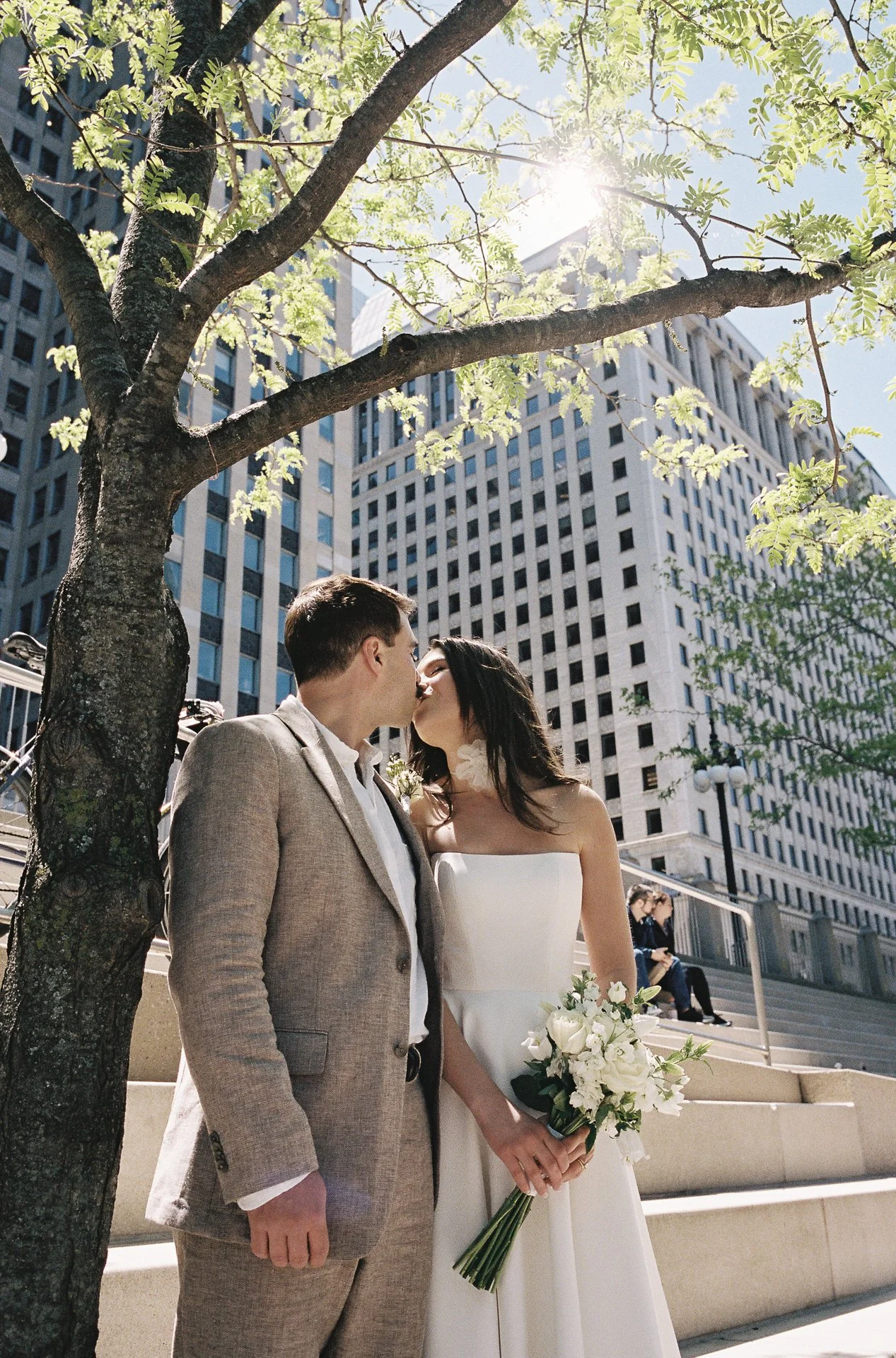 A newlywed couple sharing a kiss under a tree in an urban setting, with tall buildings in the background, sunlight filtering through the leaves, and the bride holding a bouquet of white flowers.
