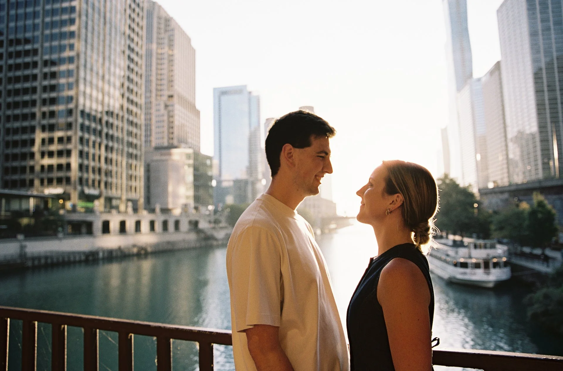 A young couple stands close together on a bridge by a river, with tall city buildings in the background, during sunset.