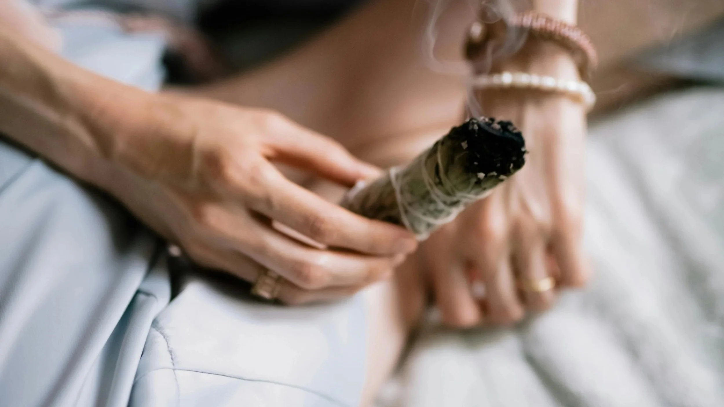 Close-up of a person holding a sage smudge stick, lying on a bed with white sheets, wearing rings and jewelry.