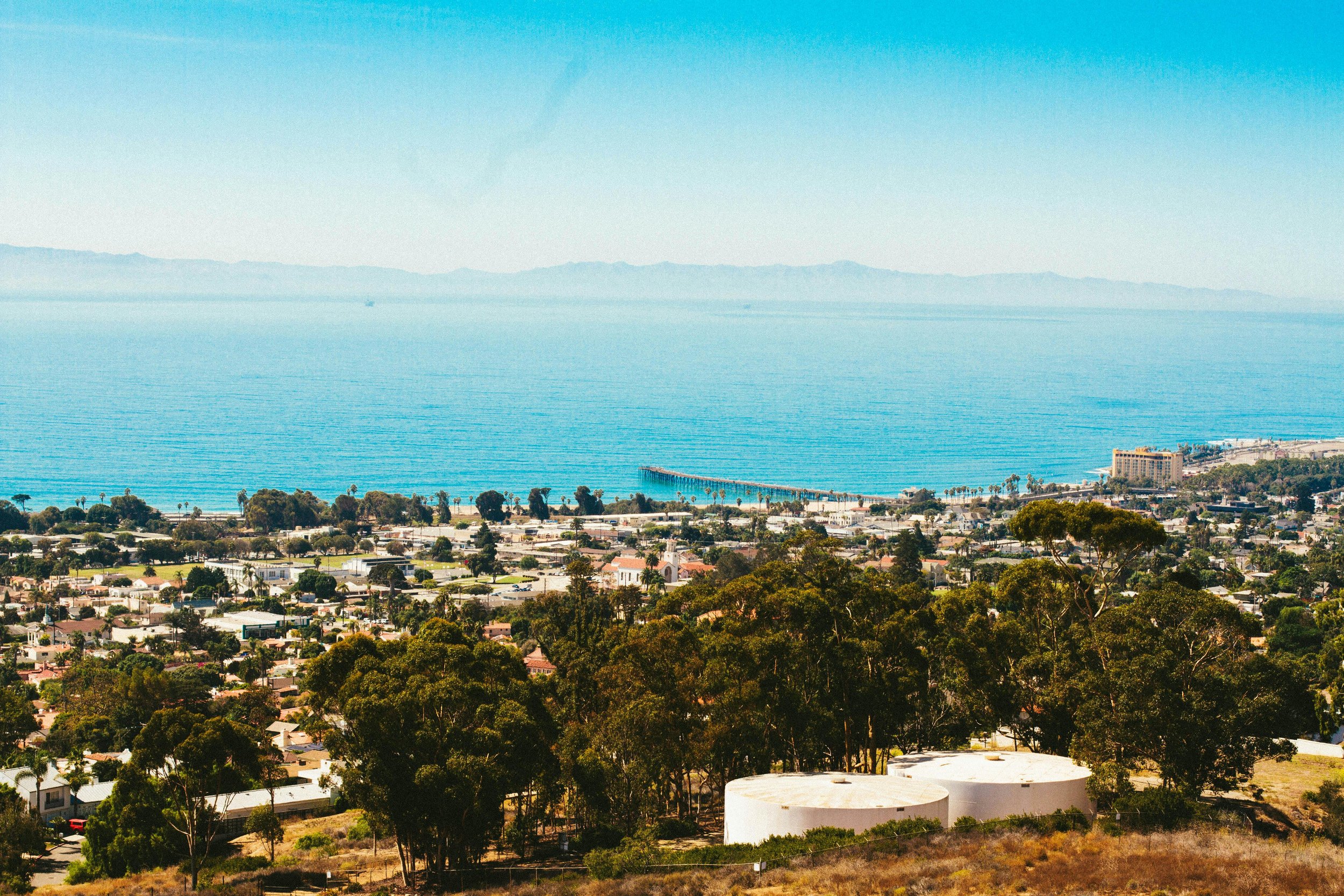 A scenic view of a coastal town with a beach, pier, and ocean, surrounded by trees and residential areas under a clear blue sky.