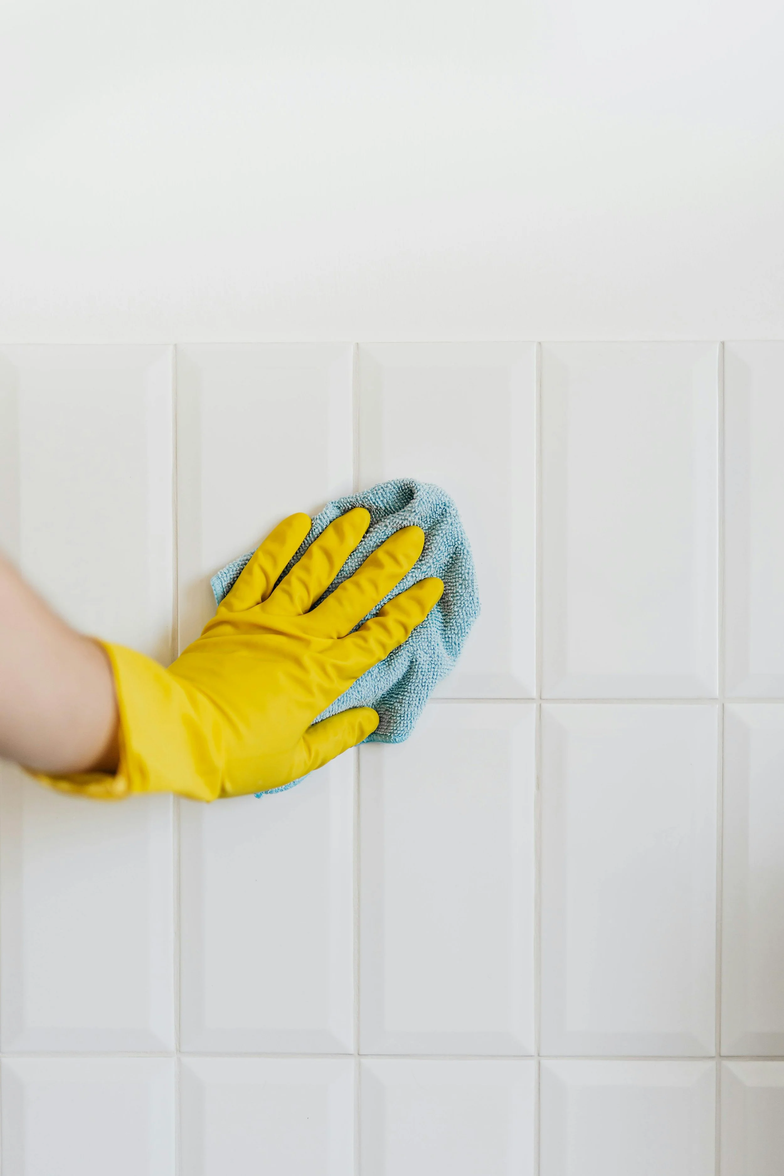 Person wearing yellow rubber gloves cleaning white tiled wall with a blue cloth.
