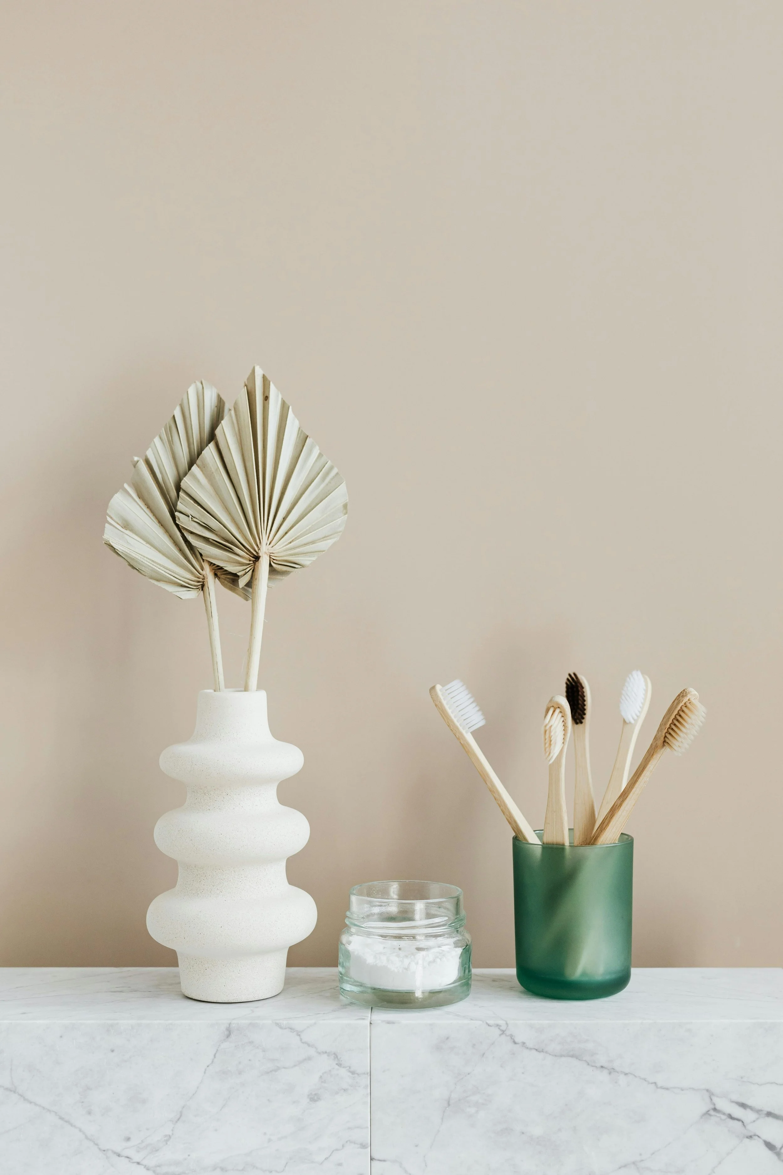 A white textured vase holding dried palm leaves, a small glass jar, and a frosted green glass container holding bamboo toothbrushes.