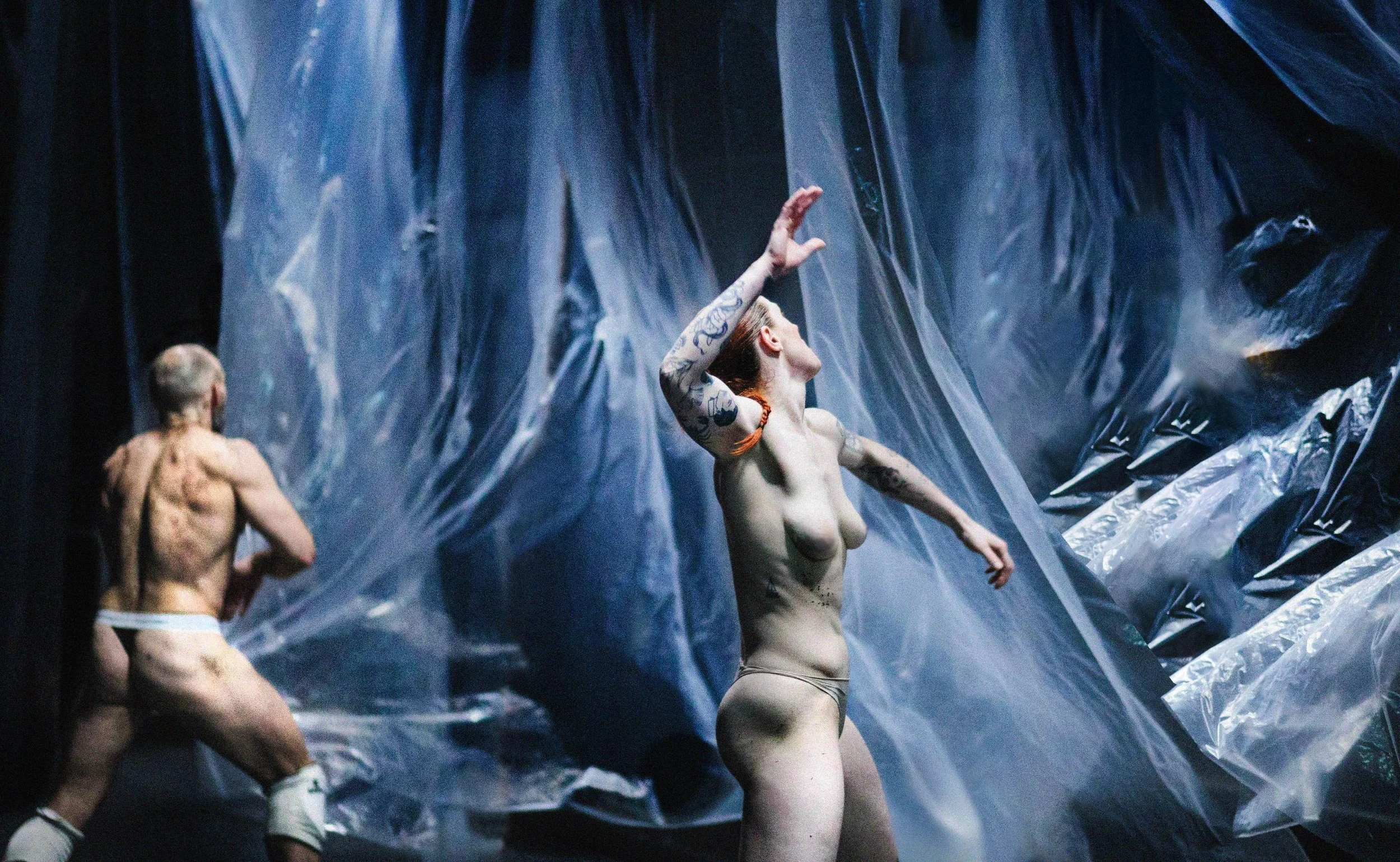 Two tattooed performers, a man and a woman, are on stage with plastic sheets behind them. The woman is posing with her arm raised and head tilted back, while the man is in motion facing away from the camera.