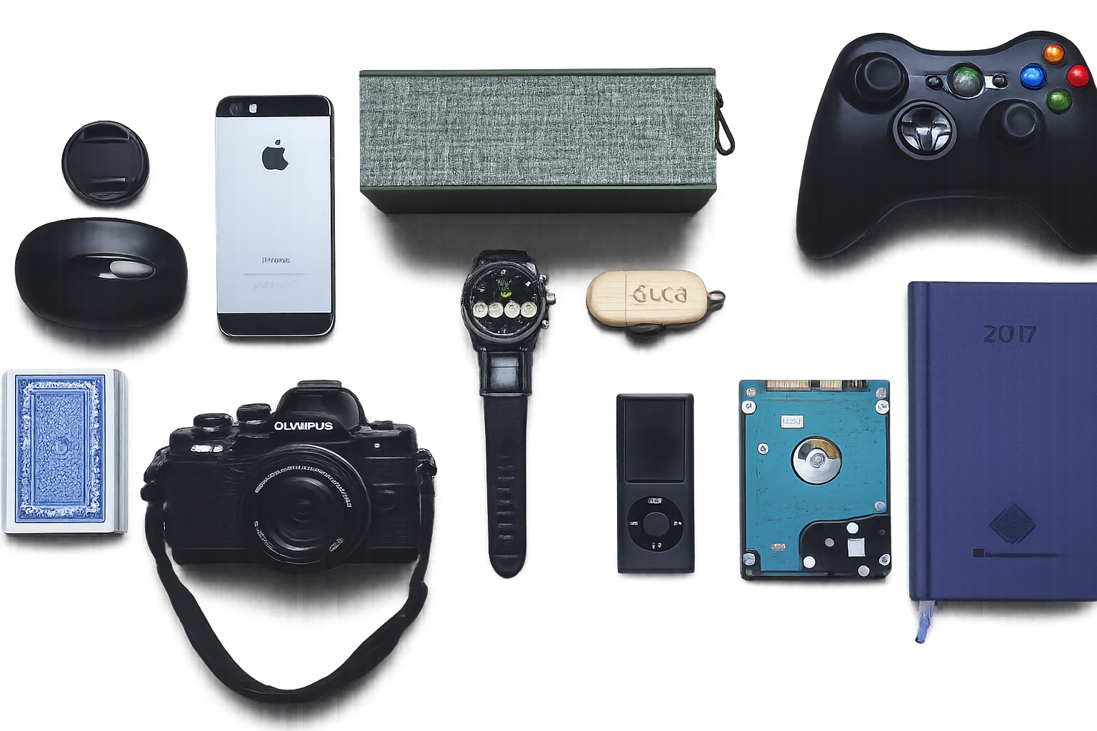A collection of personal items on a gray surface, including a smartphone, camera, smartwatch, portable speaker, external hard drive, gaming controller, watch, deck of playing cards, USB flash drive, and a blue photo album.
