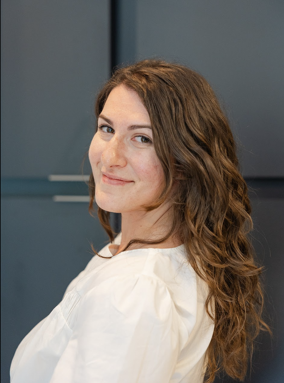 A woman with wavy brown hair, light skin, and wearing a white blouse, smiling slightly and looking at the camera against a dark background.