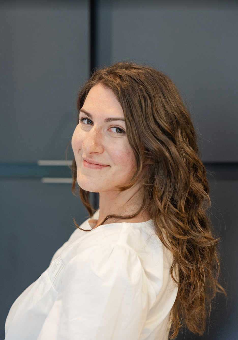 A woman with wavy brown hair wearing a white top, looking over her shoulder, smiling slightly, standing against a dark background.