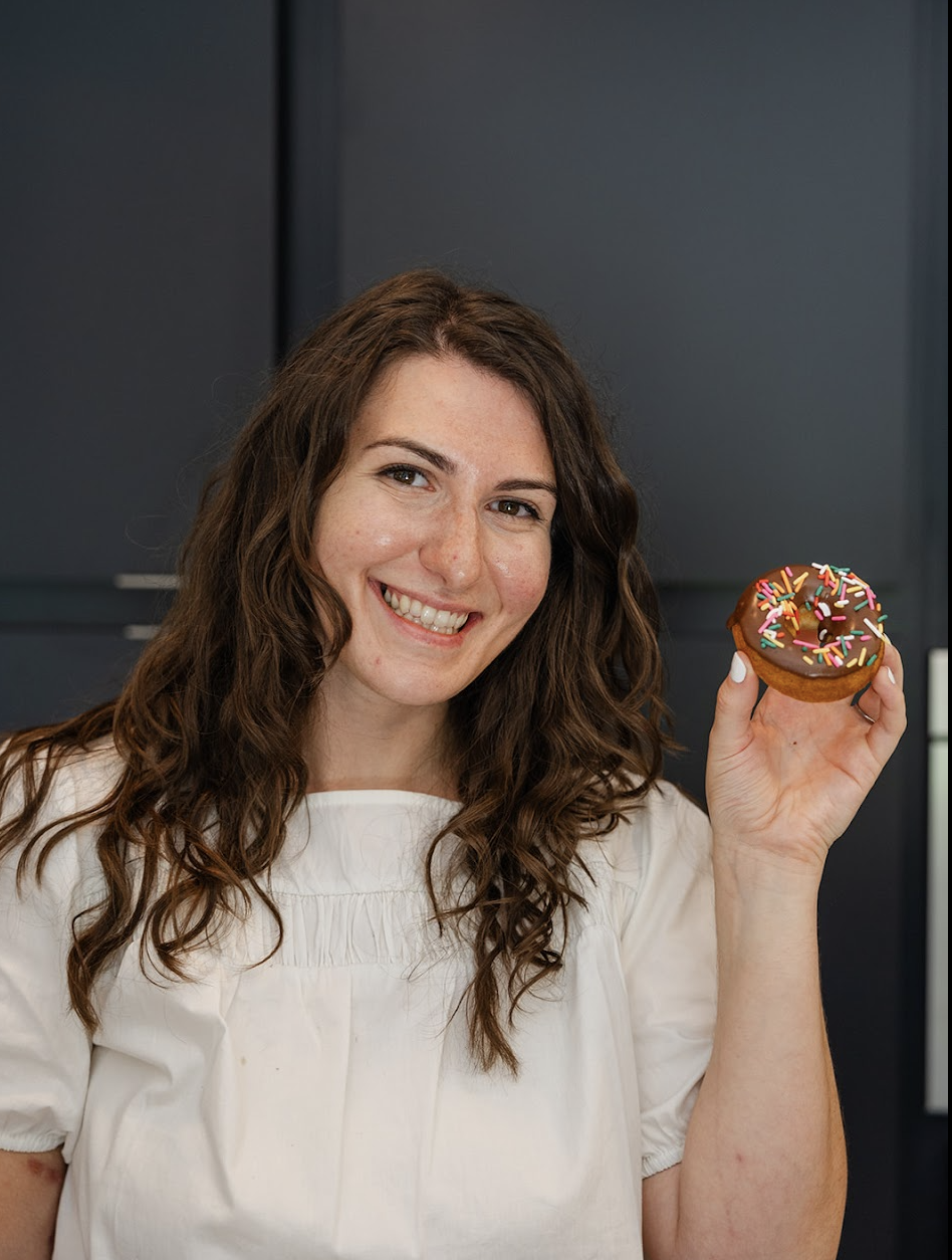 A smiling woman with long, wavy brown hair holding a chocolate donut with colorful sprinkles in a modern kitchen.