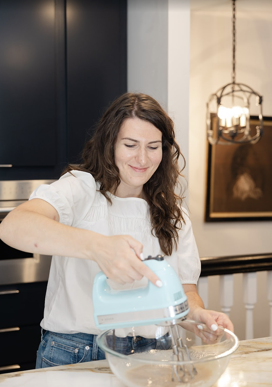 A woman with long curly brown hair and a white blouse is mixing ingredients with an electric hand mixer in a glass bowl in a modern kitchen.