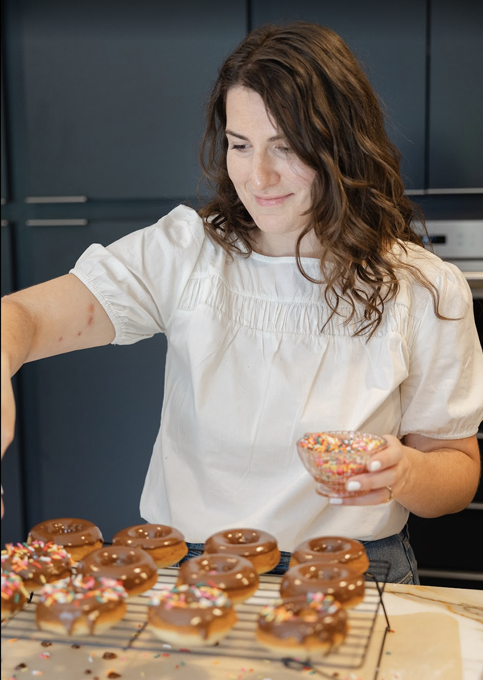 A woman decorating chocolate glazed donuts with colorful sprinkles in a kitchen.