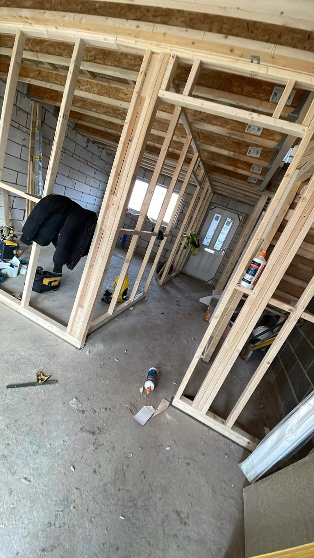 Interior of a house under construction with wooden framing, tools, and construction materials scattered around. A door and windows are visible in the background.