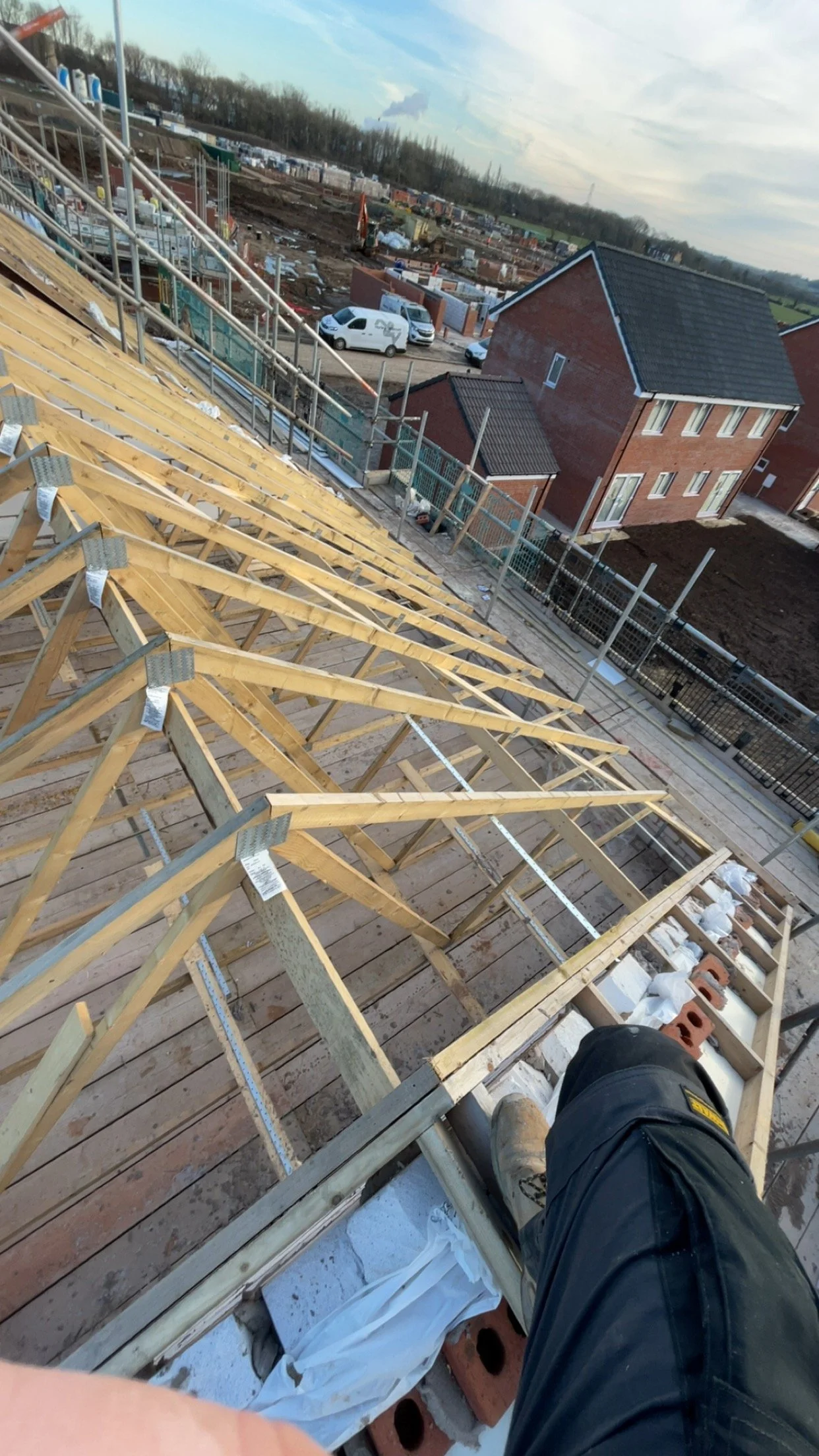 View from a construction site showing the roof framework of a building under construction with wooden trusses and bricks, with a residential area and cloudy sky in the background.