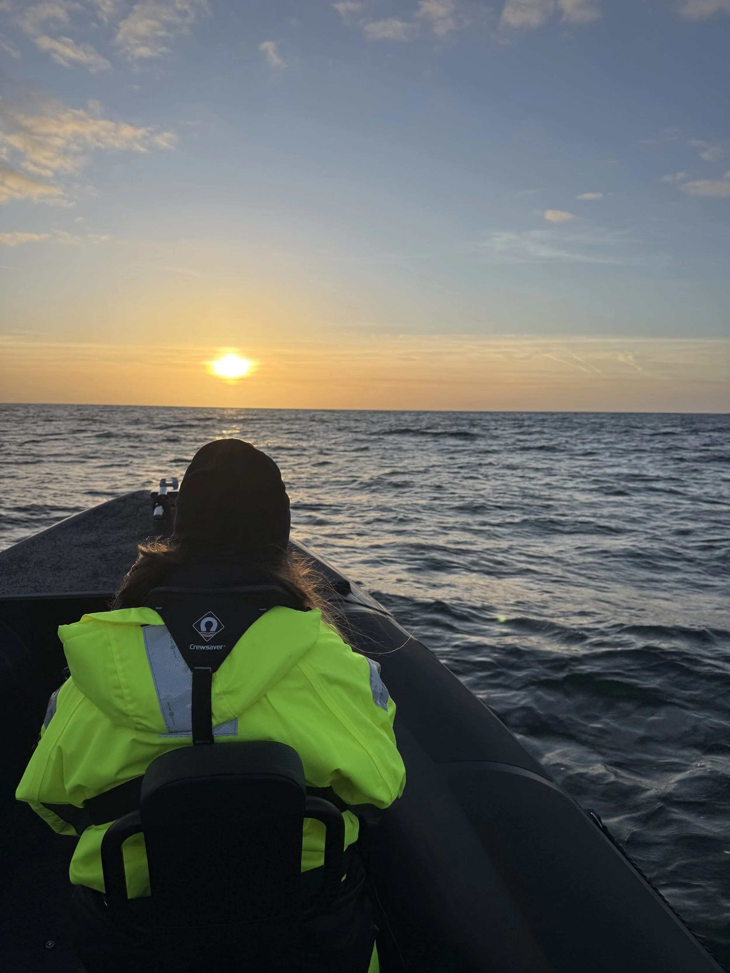 A person wearing a bright yellow safety jacket on a boat, facing the sunset over the ocean with clouds in the sky.