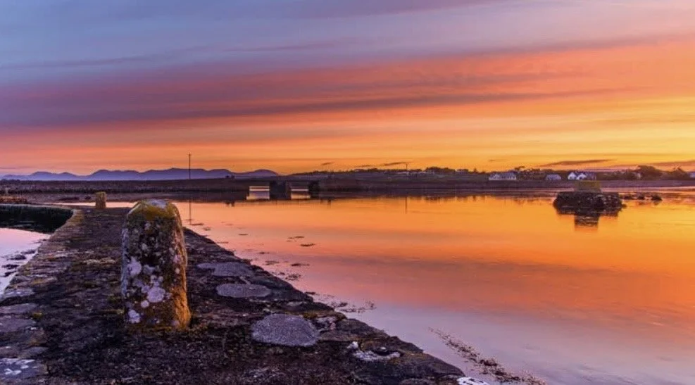 Sunset over a calm body of water with a stone pier extending into it, and buildings and mountains on the horizon.