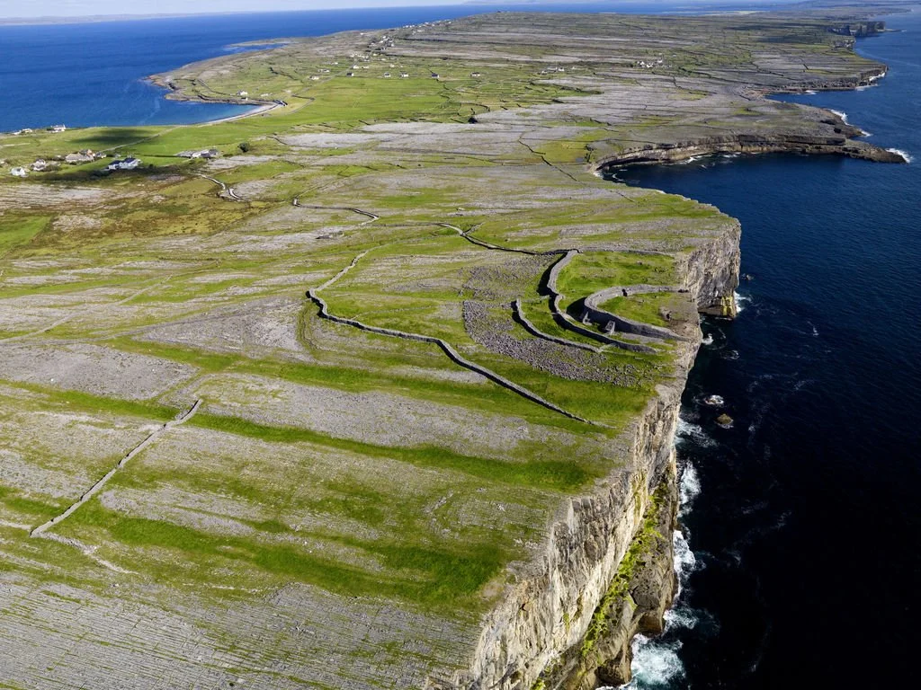 Aerial view of a rugged coastline with green fields, stone walls, and a cliff edge over the ocean in Ireland.