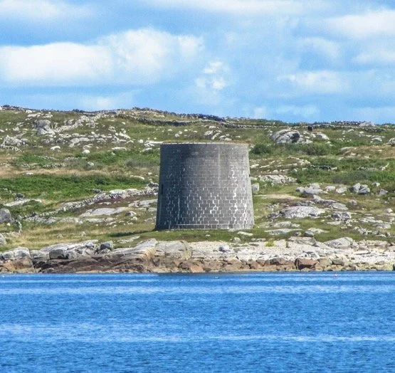 A cylindrical stone structure on a grassy hillside near a body of water with a cloudy sky in the background.