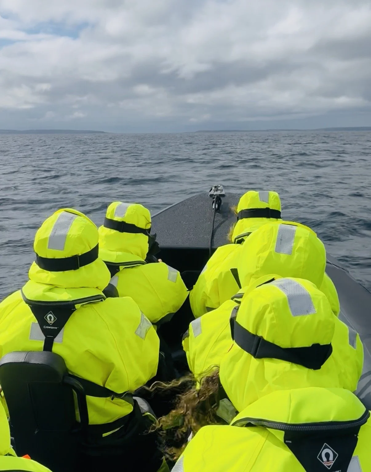 A group of people in bright yellow life jackets and yellow raincoats sitting on a boat, facing the water with overcast skies.