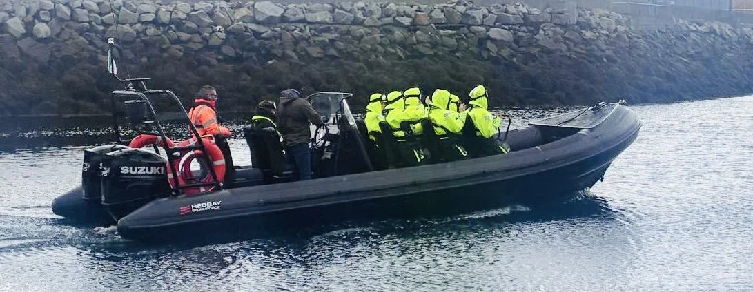 A black rigid inflatable boat with a Suzuki engine, carrying a group of law enforcement officers wearing bright yellow jackets and helmets, on a body of water near a rocky shoreline.