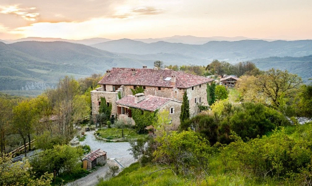 A stone house with a red-tiled roof situated on a lush green hillside, surrounded by trees and overlooking distant rolling mountains at sunset.