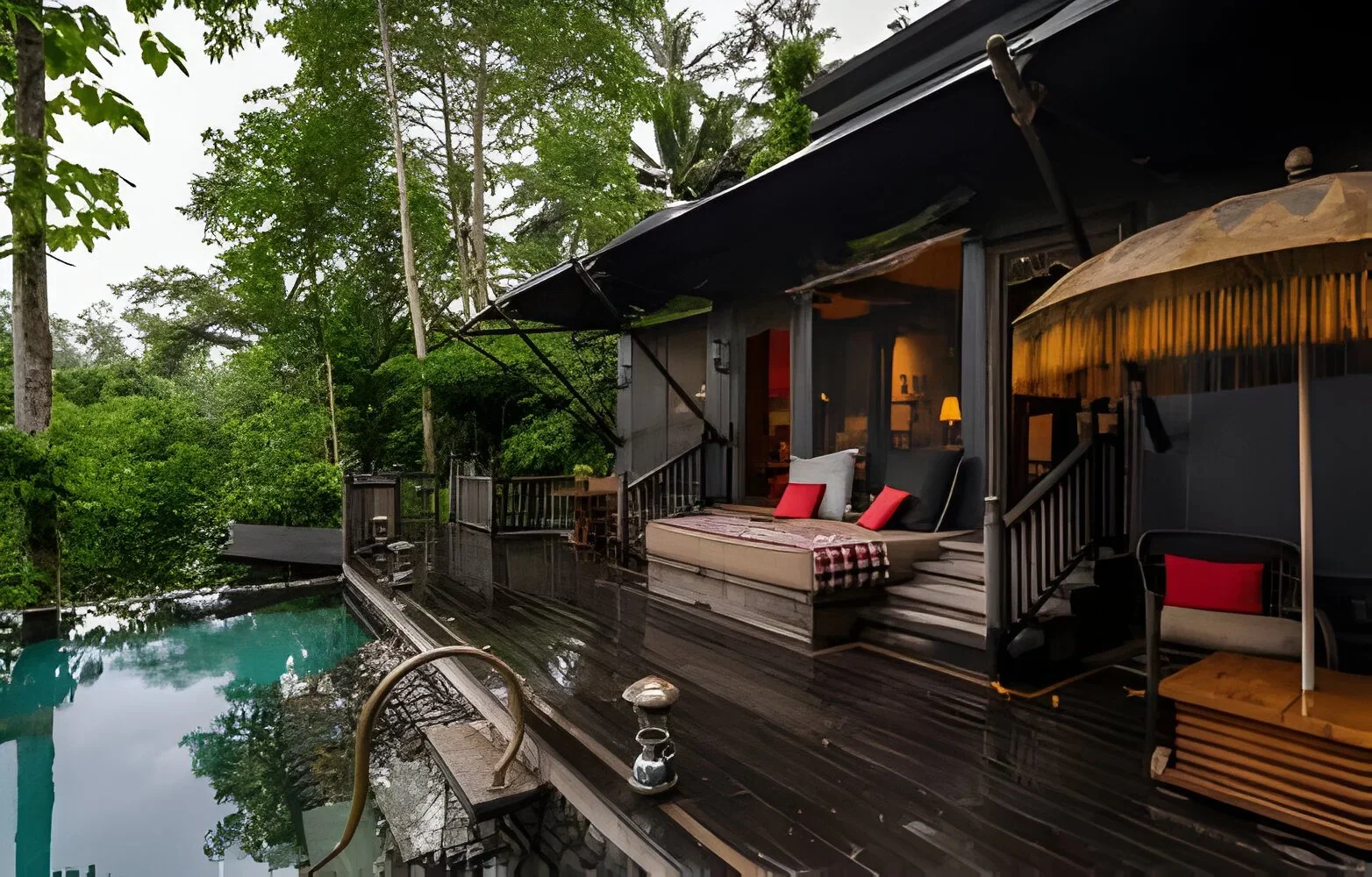 Outdoor deck of a house with a pool, surrounded by trees and greenery, featuring seating with cushions and an umbrella under an overcast sky.
