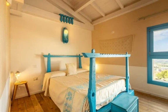 A bedroom with white walls and a pitched ceiling, featuring a bed with a blue wooden canopy frame and matching blue suitcase at the foot, a small wooden side table with a lamp, a large window with blue trim, and a woven wall hanging.
