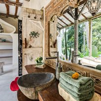 Interior of a rustic home with a large window, a round black sink, shelves with potted plants, and a cozy seating area outside