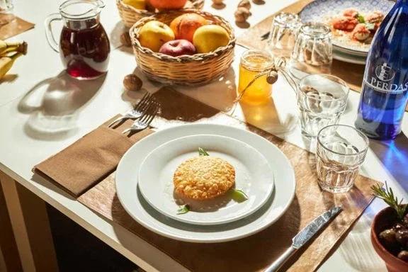 A table set with a plate of breaded fried food garnished with greens, a basket of apples, a pitcher of juice, glasses, and a bottle of water, with additional plates and glasses in the background.
