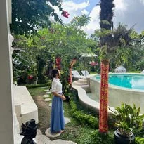 A woman standing near a swimming pool surrounded by lush green trees and plants.