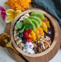 A wooden bowl filled with assorted fresh fruit slices including kiwi, mango, strawberries, and blueberries, along with a variety of granola and shredded coconut, garnished with flowers.