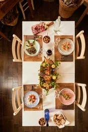 A long dining table set for a meal, with chairs on each side, decorated with a floral centerpiece and various dishes and drinks.