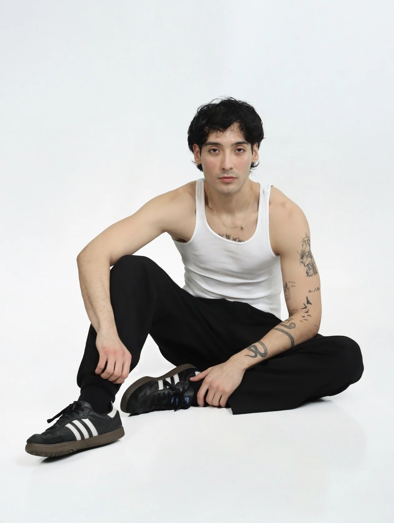 Jordan Campos wearing a white tank top and black pants, sitting on the floor against a plain white background, with tattoos on his arms and black sneakers with white stripes showing commercial lifestyle and commercial modeling look.