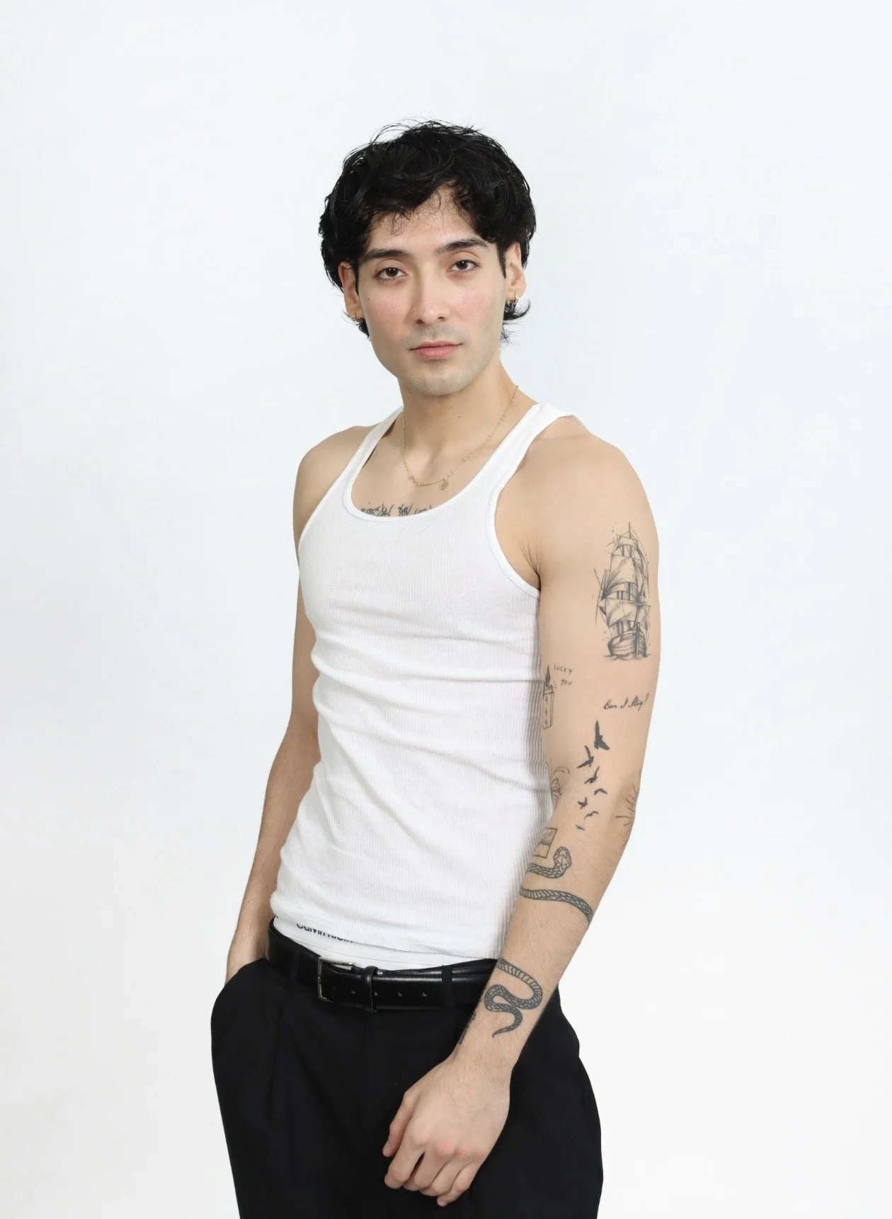 Jordan Campos with black hair and tattoos on his left arm, wearing a white tank top and black pants, standing against a plain white background showing in a fit lifestyle look.