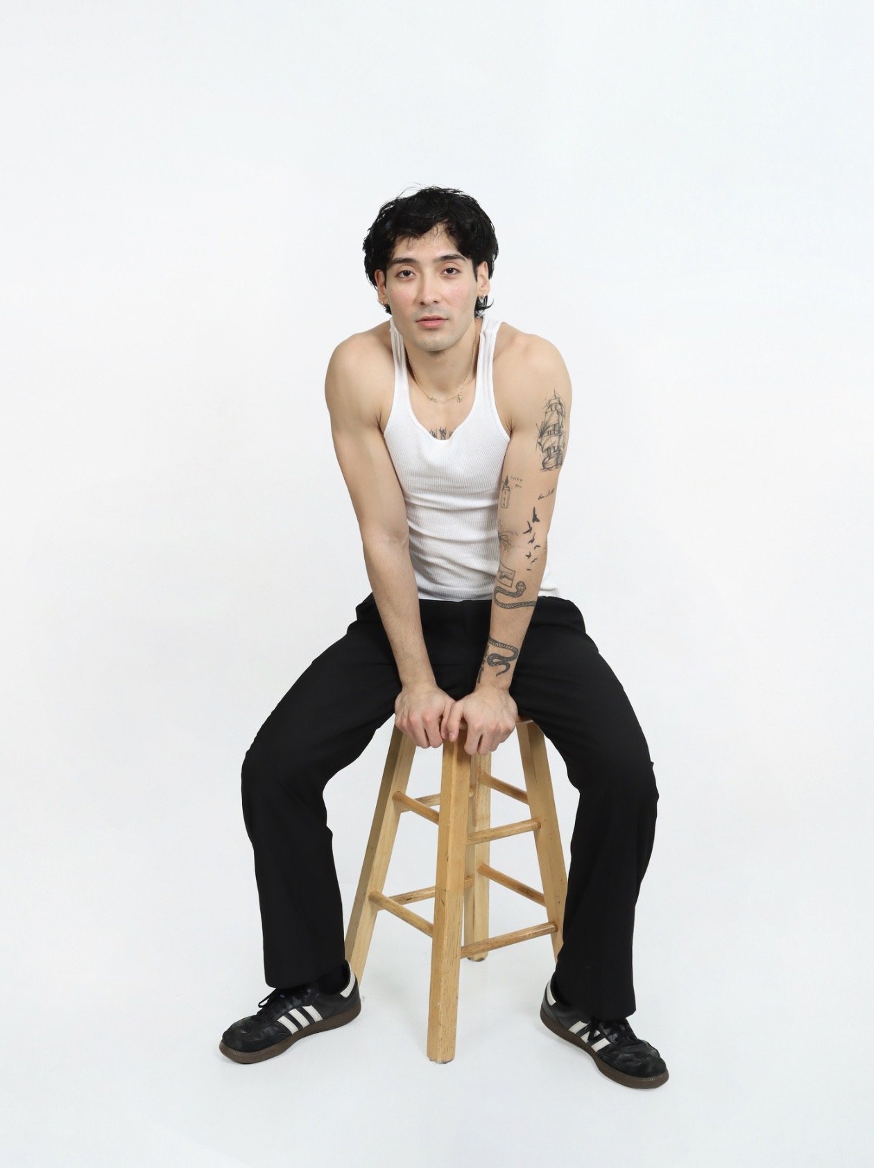 Jordan Campos with black hair, white tank top, black pants, and black sneakers sits on a wooden stool against a plain white background, looking directly at the camera giving modern commercial and fitness lifestyle modeling look.
