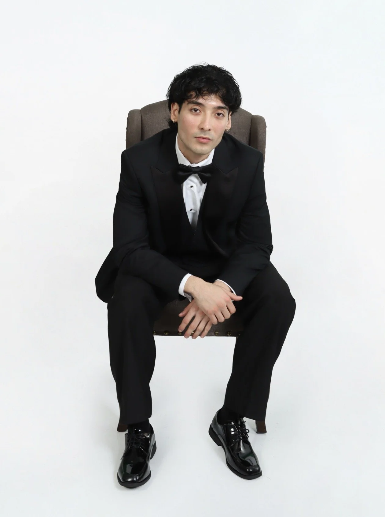 Jordan Campos in formal wear attire, black tuxedo with a bowtie, black dress shoes, sitting on a brown chair against a plain white background.