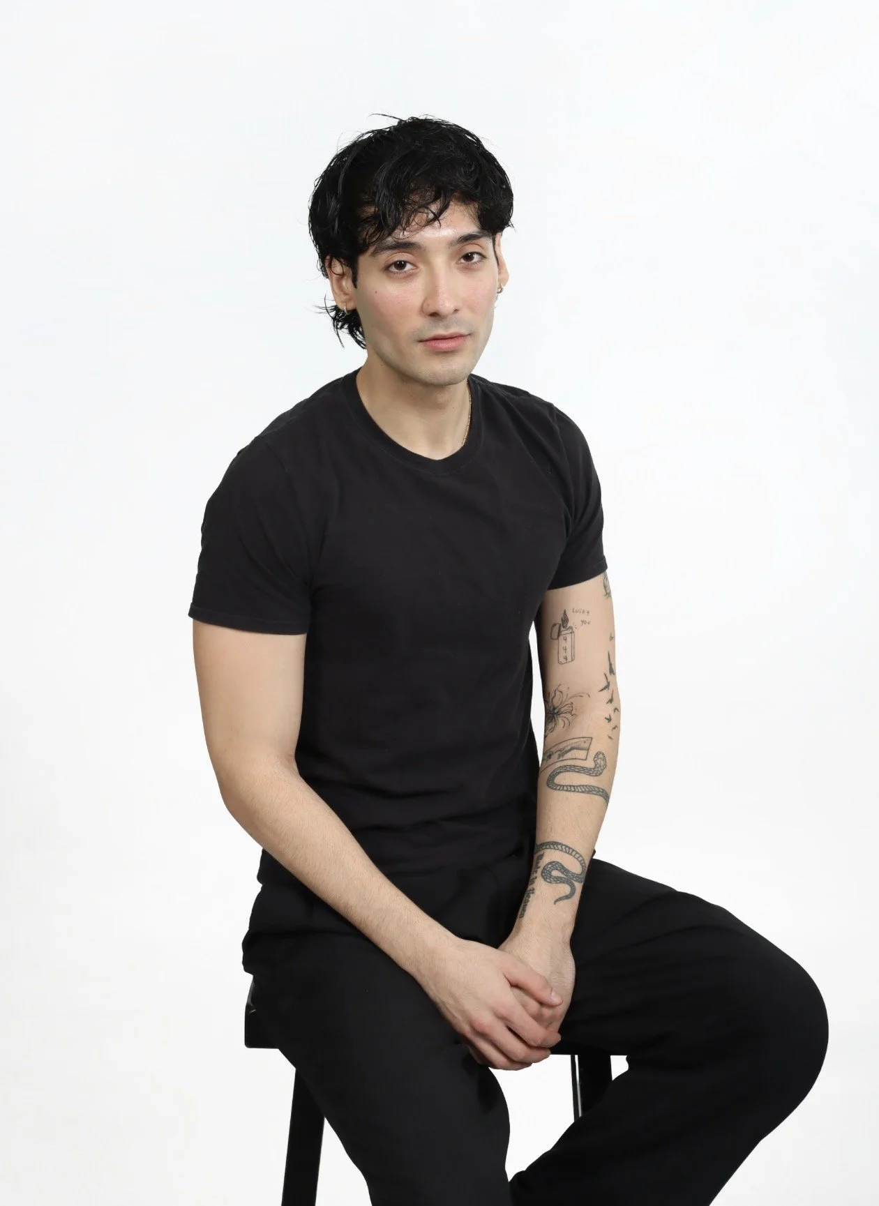 Jordan Campos with black hair, wearing a black t-shirt, sitting on a stool against a plain white background showcasing niche and natural expression.